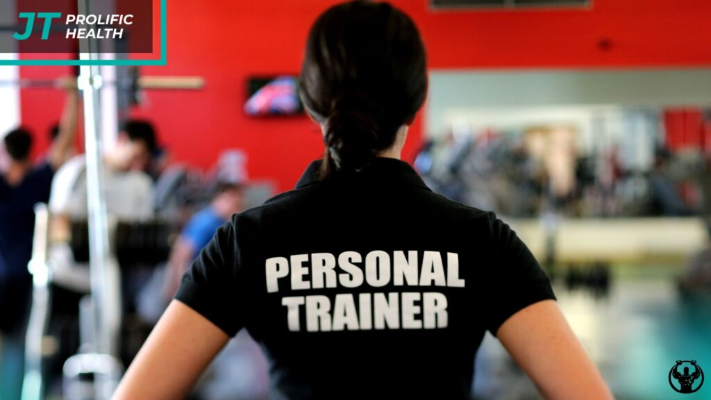 Personal trainer in a gym setting, wearing a shirt labeled "PERSONAL TRAINER," with fitness equipment and individuals exercising in the background, emphasizing professional coaching and fitness guidance at Prolific Health.