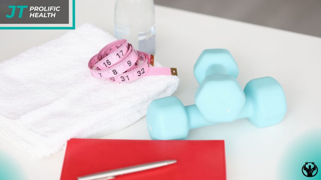 Blue dumbbell, measuring tape, towel, water bottle, and notebook on a table, symbolizing personal training and fitness planning at Prolific Health.