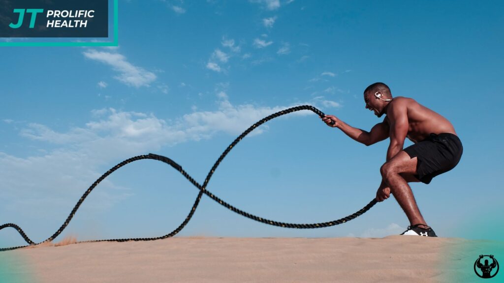 Man performing battle rope exercises on sandy terrain, emphasizing high-energy training and fitness progression, with Prolific Health branding visible.